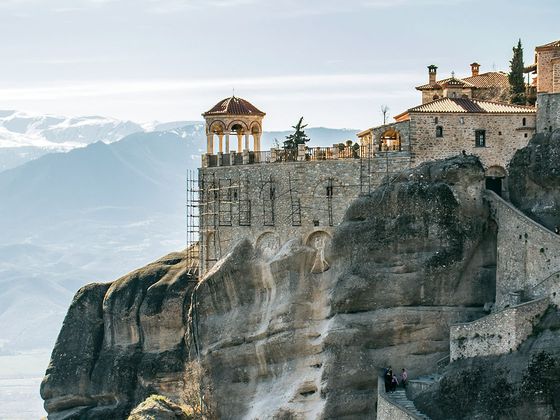 Historic monastery perched on a steep rock formation with distant snow-capped mountains and a clear sky in the background.