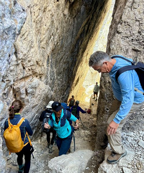 Hikers navigate a narrow, rocky canyon passage, with some climbing and others using trekking poles, surrounded by steep stone walls.