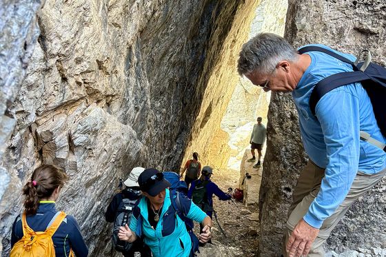 Hikers navigate a narrow, rocky canyon passage, with some climbing and others using trekking poles, surrounded by steep stone walls.