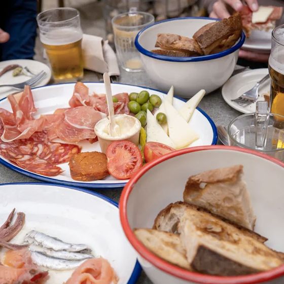 Assorted cold cuts, cheese, olives, and bread on plates with drinks in the background, set on a table in an outdoor dining area.