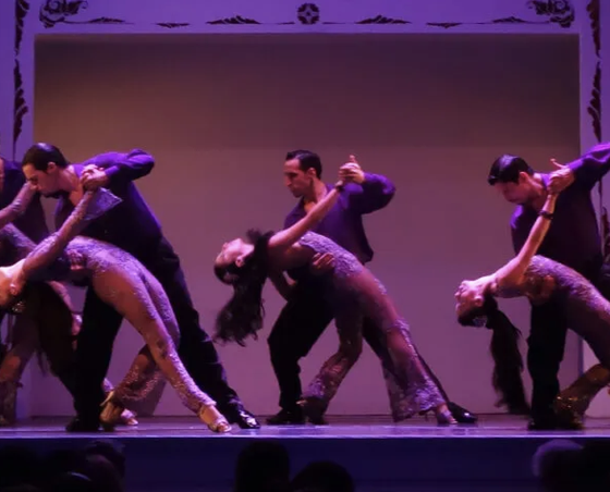 Three couples perform a synchronized dance on stage, wearing elegant costumes under purple lighting.