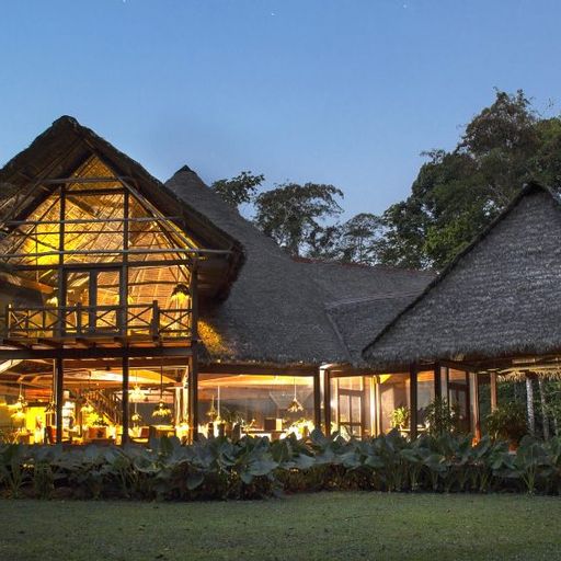 A well-lit, rustic lodge with a thatched roof surrounded by lush greenery under a twilight sky.
