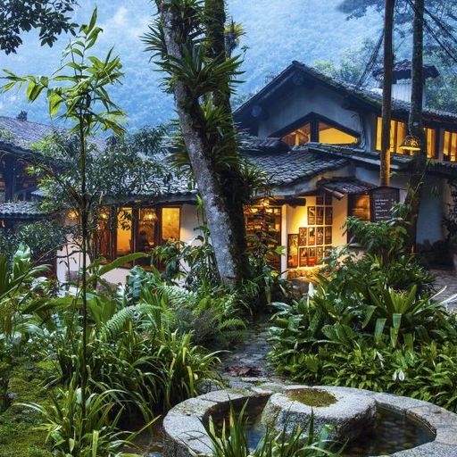 A lush, green garden surrounds a cozy house with warm, glowing windows, set against a backdrop of misty mountains and tall trees.