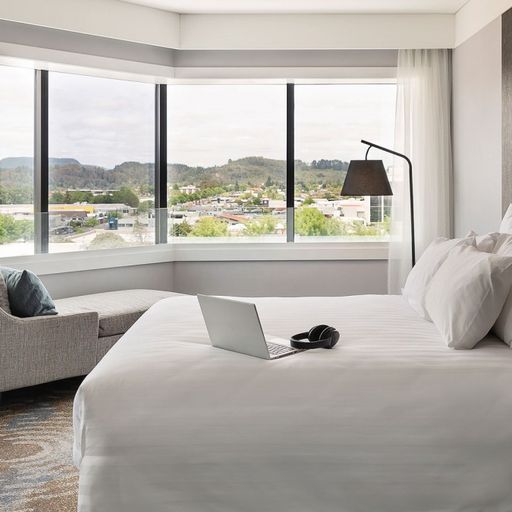Modern hotel room with a large bed, laptop, and headphones. A chaise lounge by a wide window offers a view of a cityscape and hills.