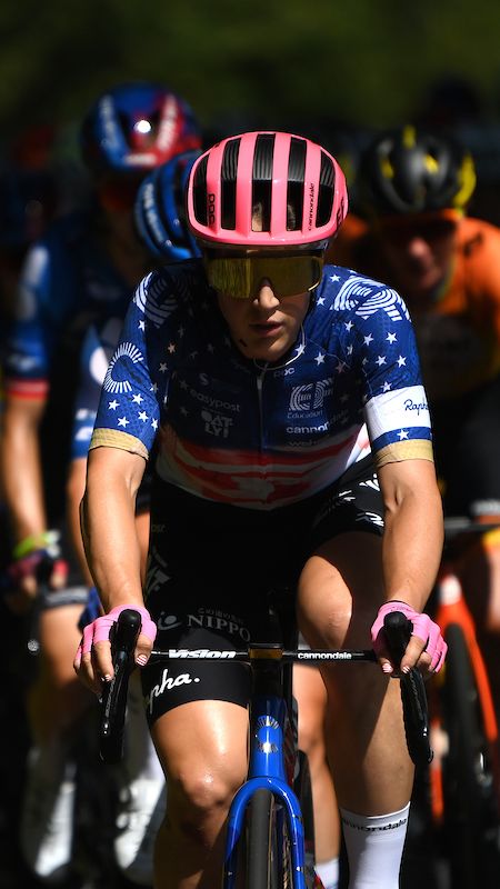 Cyclist in a blue and star-patterned jersey leads a dense pack during a race, surrounded by other cyclists in colorful helmets and jerseys.