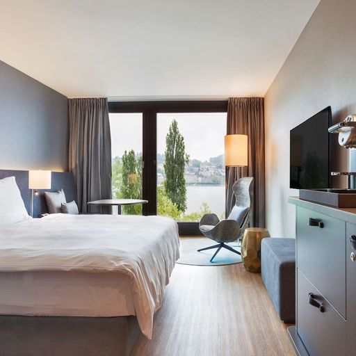 Modern hotel room with a large bed, TV, coffee maker, and a window view of trees and water. Neutral tones and minimalist decor.