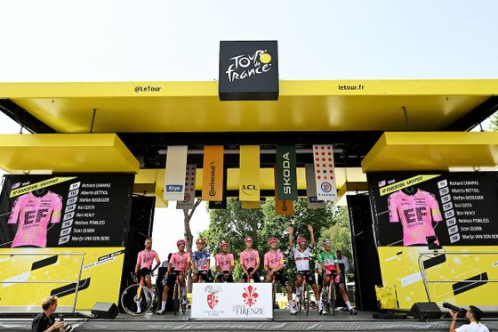 Cycling team in pink jerseys on a yellow stage at a Tour de France event, with sponsor banners and audience in the background.