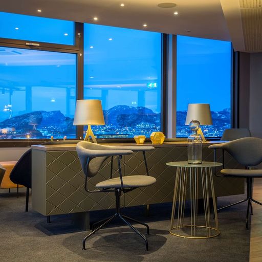 Modern lounge with stylish chairs and tables, large windows revealing a cityscape and mountains under a twilight sky. Warm indoor lighting.