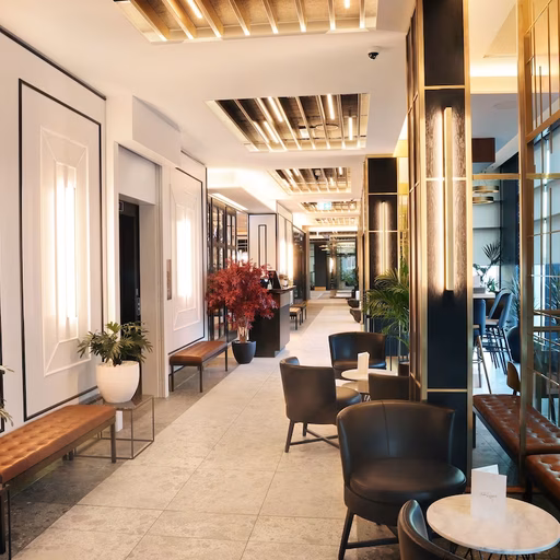 Modern hallway or lobby with light-colored walls, vertical lighting fixtures, brown leather benches, black armchairs, potted plants, and wooden ceiling panels, creating a sophisticated and spacious ambiance.