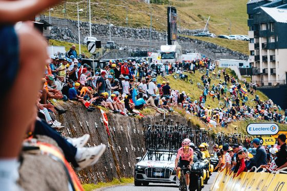 Cyclists in a race climb a mountain road lined with spectators. Support vehicles follow, and a building is visible in the background.