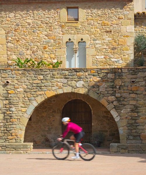 Cyclist in red jacket rides past historic stone building with arches and windows under a clear blue sky.