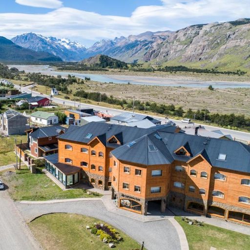 Aerial view of a large wooden building with a dark roof, surrounded by a scenic landscape of mountains, river, and small houses.