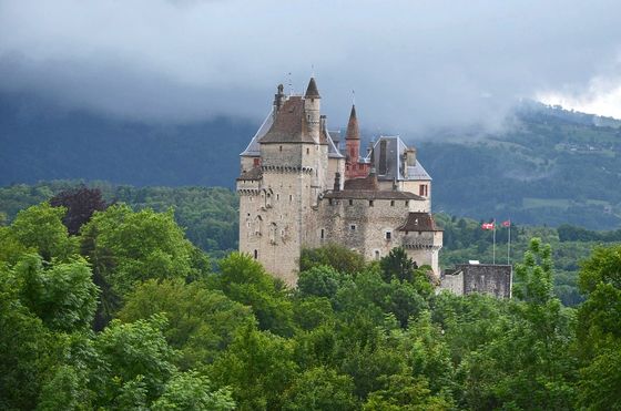 A medieval castle with towers and turrets surrounded by lush green trees, set against a backdrop of misty mountains and cloudy sky.