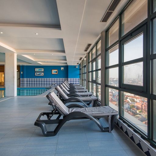 Indoor pool area with several lounge chairs facing large windows offering a city view. Blue walls and tiled accents create a modern atmosphere.