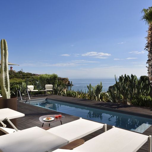 Poolside view with white loungers, a small pool, and lush greenery overlooking the ocean under a clear blue sky.