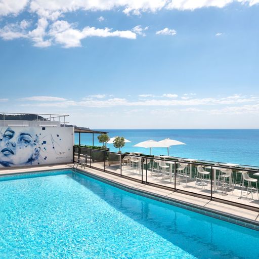 Rooftop pool with lounge chairs, a mural of a face on the wall, and a view of the ocean under a clear blue sky.