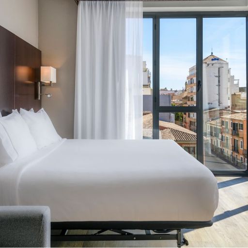 Modern hotel room with a large bed, floor-to-ceiling windows, and a city view. White curtains and wooden accents create a bright, cozy atmosphere.