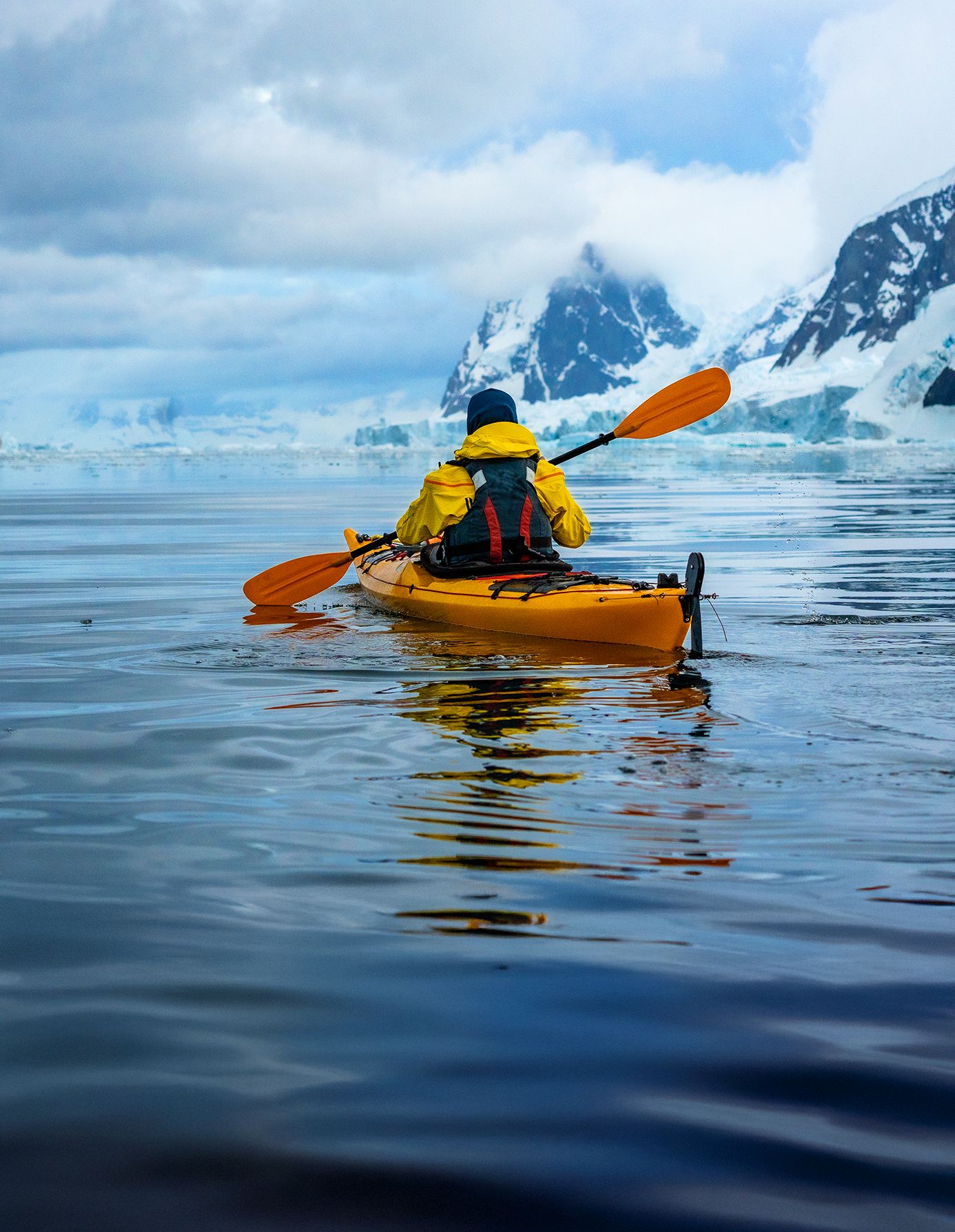 Person in a yellow kayak paddling on calm water, surrounded by snow-covered mountains under a cloudy sky.