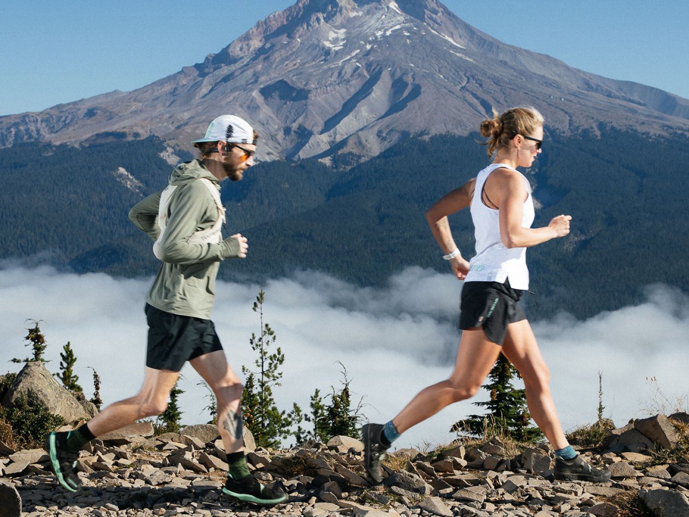 Two people trail running on a rocky path with a majestic mountain and cloud-filled sky in the background.