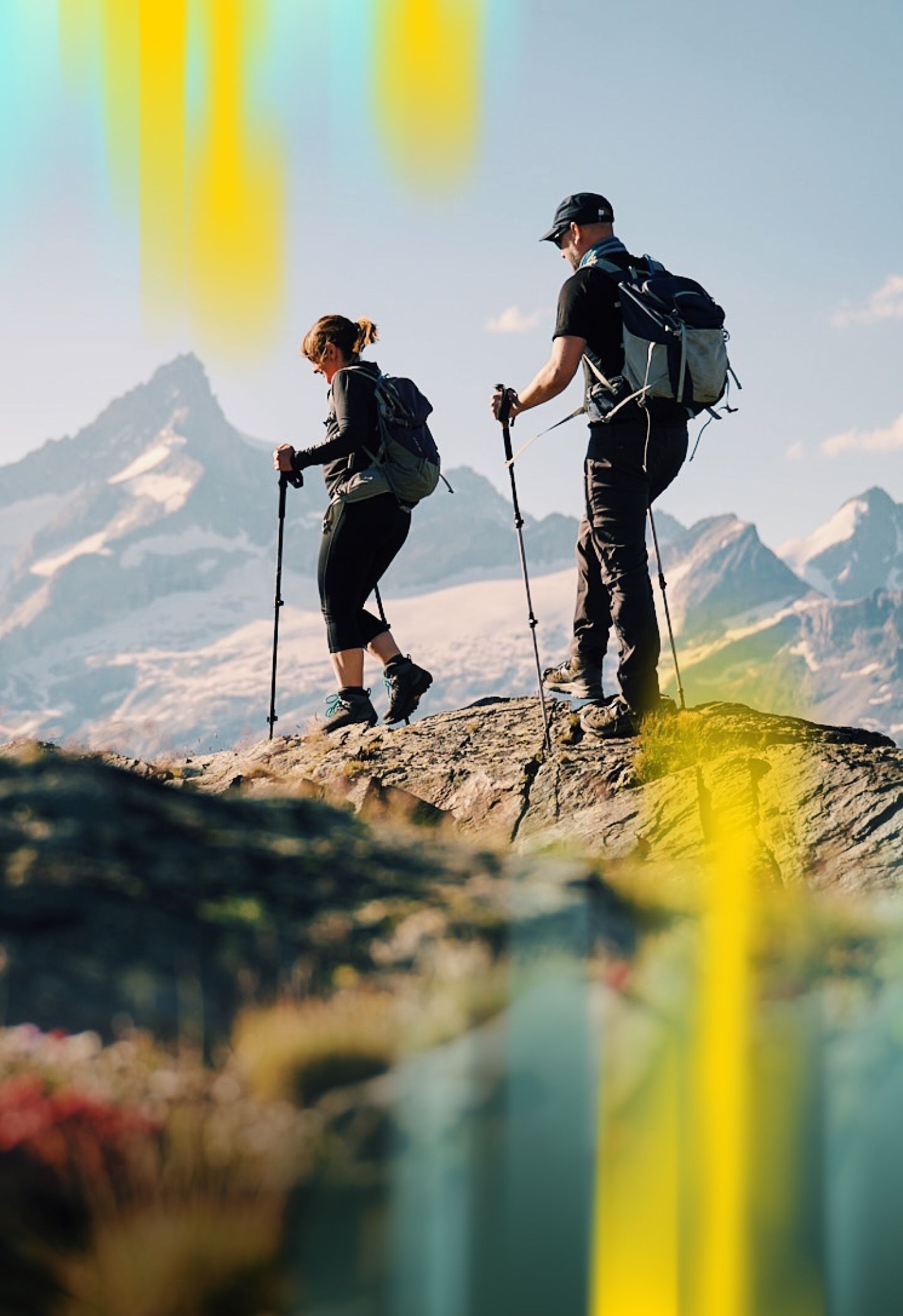 Two hikers with backpacks and trekking poles walk on a rocky mountain trail, with snow-capped peaks in the background under a clear sky.