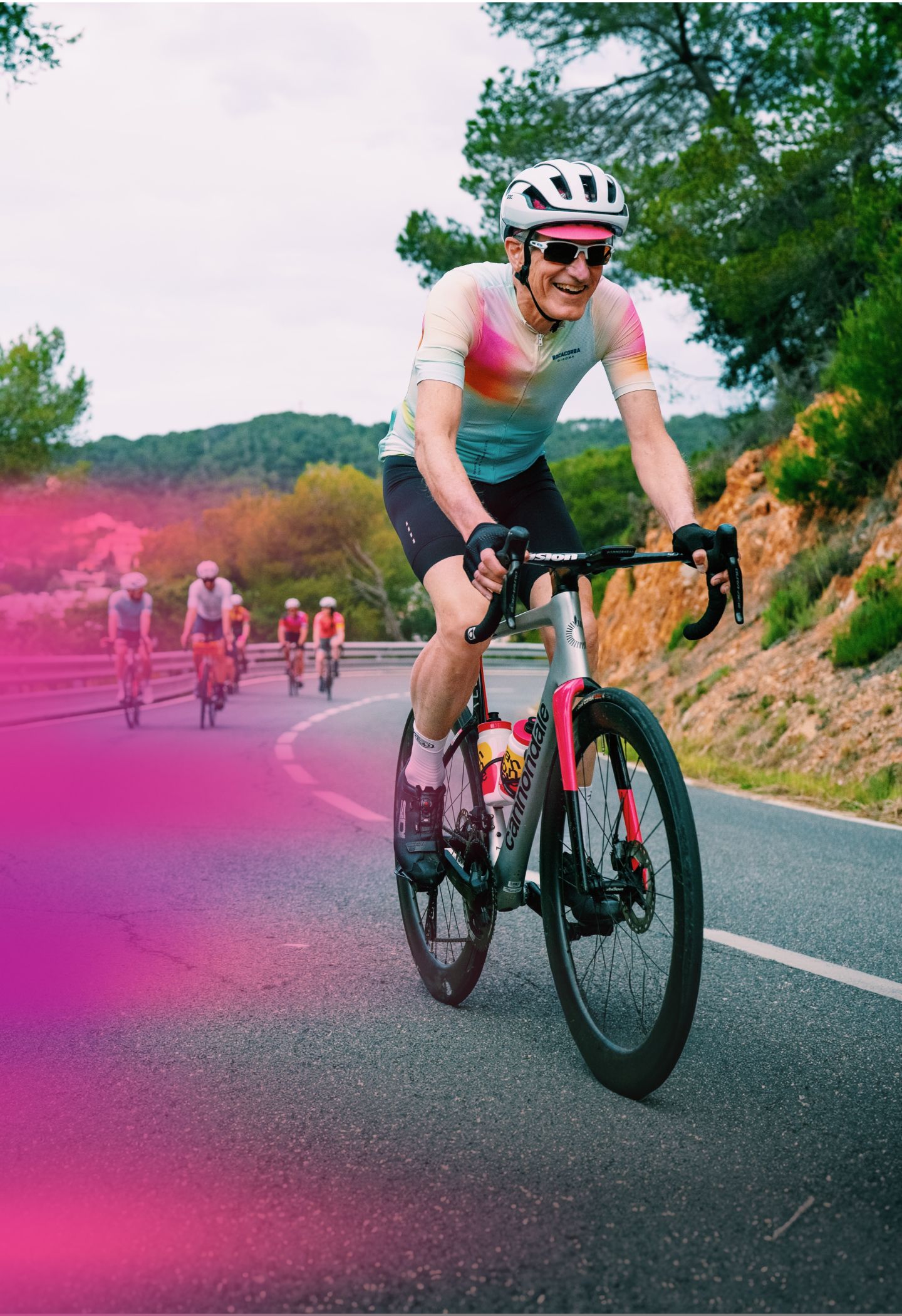 Cyclist in a colorful jersey rides uphill on a scenic road with trees, followed by a group of cyclists.