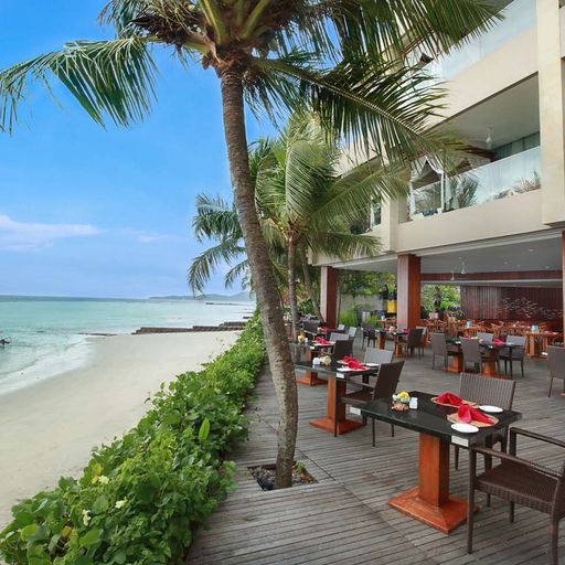 Beachfront restaurant with outdoor seating, palm trees, and a view of the ocean. Tables are set with red napkins and white plates.