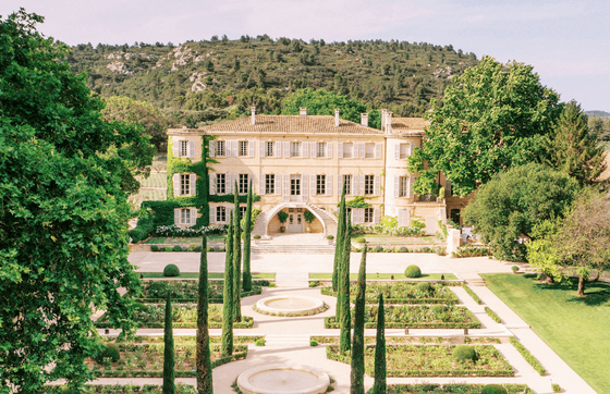 Elegant villa surrounded by manicured gardens, cypress trees, and lush greenery, set against a backdrop of rolling hills and blue sky.