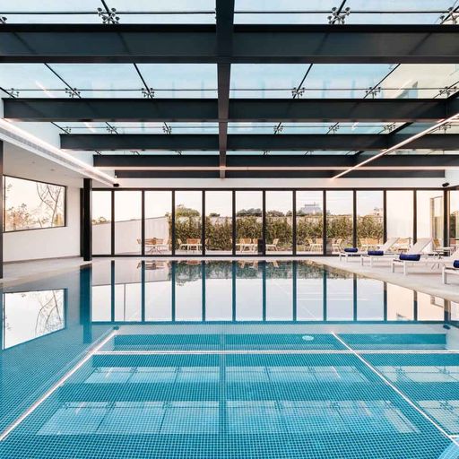 Indoor pool with clear blue water, surrounded by white lounge chairs. Large windows and a glass ceiling allow natural light to fill the space.