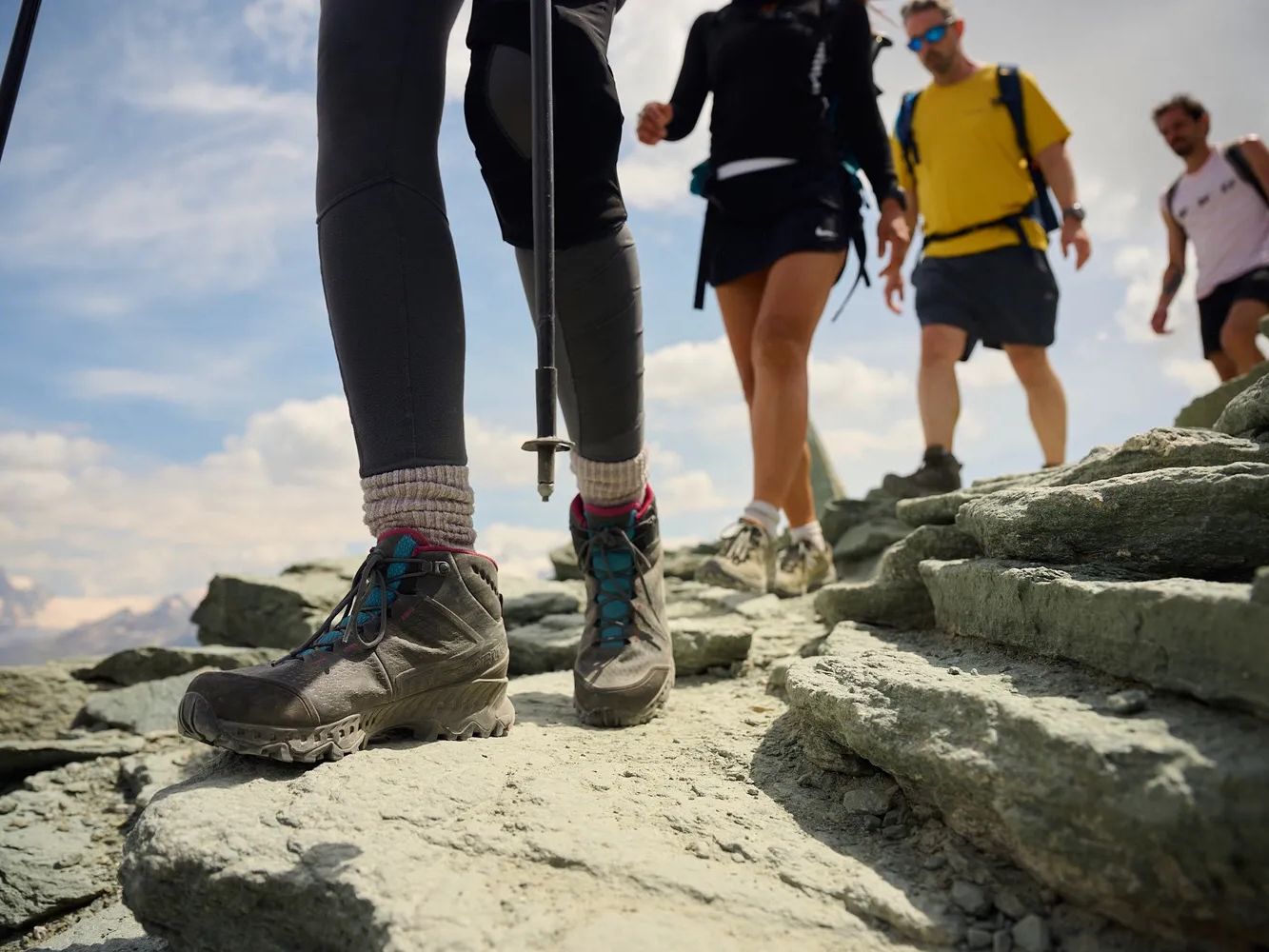 Hikers walking on a rocky mountain trail, with focus on the leading person's hiking boots and trekking pole, under a partly cloudy sky.