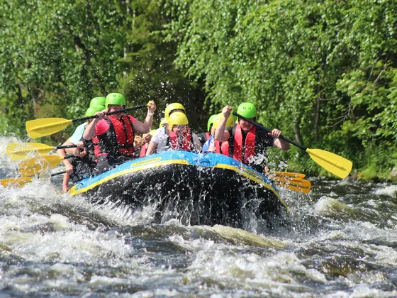 Group of people in helmets and life jackets white-water rafting on a river, surrounded by lush green trees.