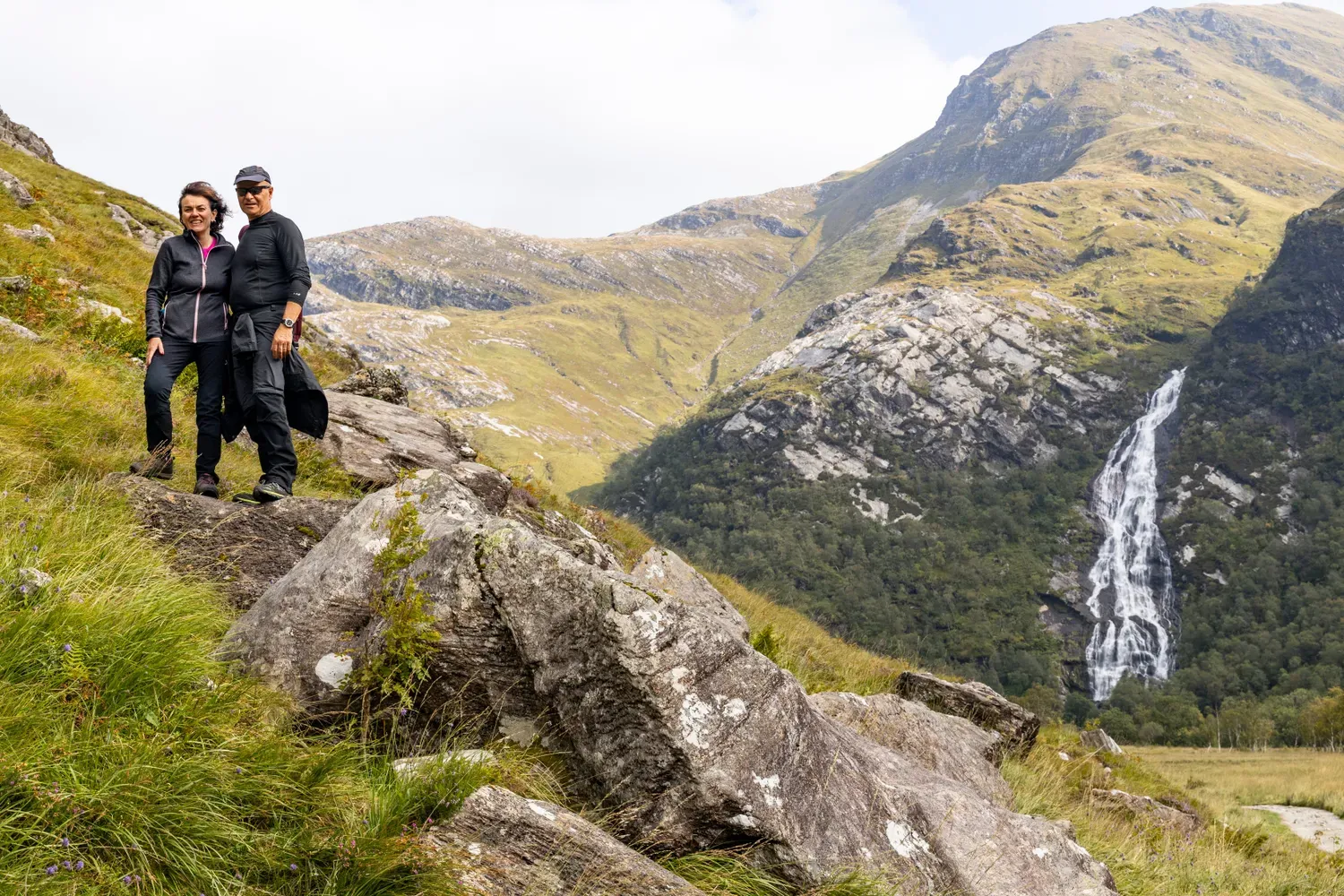 Two hikers standing on rocky terrain with a scenic waterfall and lush green mountains in the background.