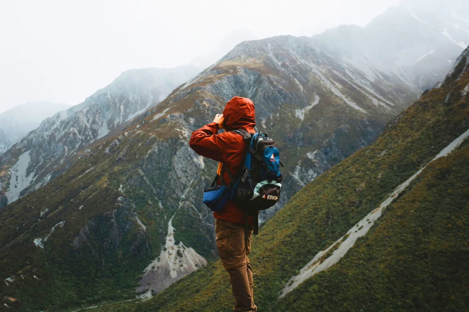 A person in an orange jacket stands on a mountain ledge, taking a photo of the misty, rugged mountain landscape with a backdrop of cloudy skies.