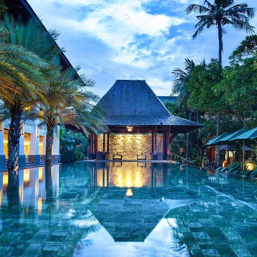 Luxurious tropical resort pool at dusk, surrounded by palm trees and lounge chairs, with a thatched-roof pavilion reflecting on the water.