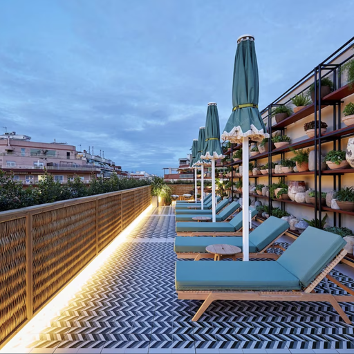Rooftop terrace with blue lounge chairs, green umbrellas, and potted plants on a patterned floor, overlooking city buildings at dusk.