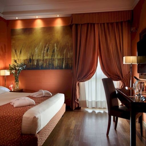 Elegant hotel room with a large bed, warm lighting, abstract wall art, desk, and window with heavy curtains. A robe and towels are on the bed.