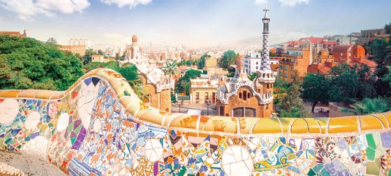 Colorful mosaic tiles adorn a curved bench overlooking the architectural structures and lush greenery of Park Güell in Barcelona.
