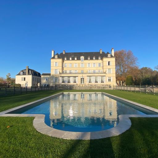 A grand chateau with a symmetrical facade is reflected in a large, clear swimming pool, surrounded by a manicured lawn and trees under a blue sky.