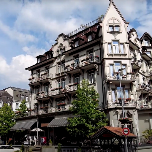 Historic European-style hotel with ornate balconies and steep roofs, surrounded by trees and a few parked cars, under a partly cloudy sky.