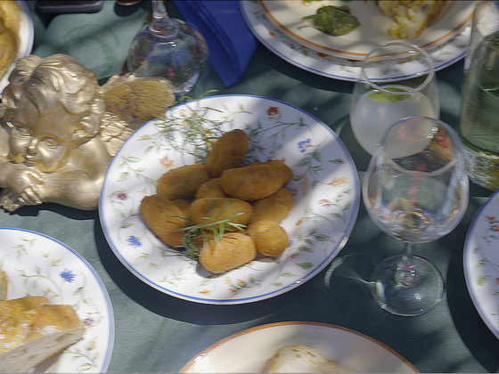 Ornate table setting with breaded appetizers on a floral plate, surrounded by wine glasses, drinks, bread, and a decorative cherub statue.