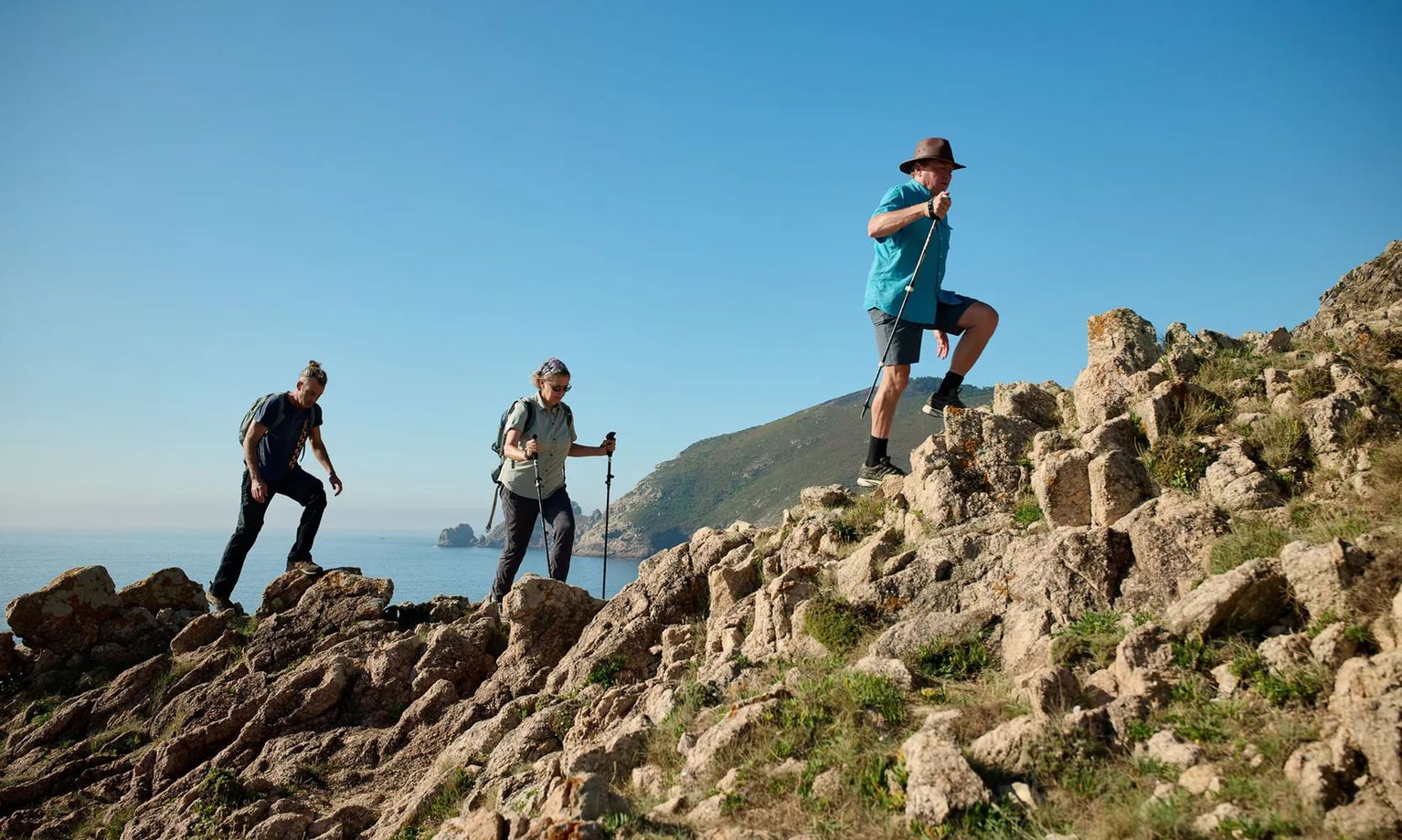 Three hikers with trekking poles climb a rocky hill under a clear blue sky, with coastal scenery in the background.