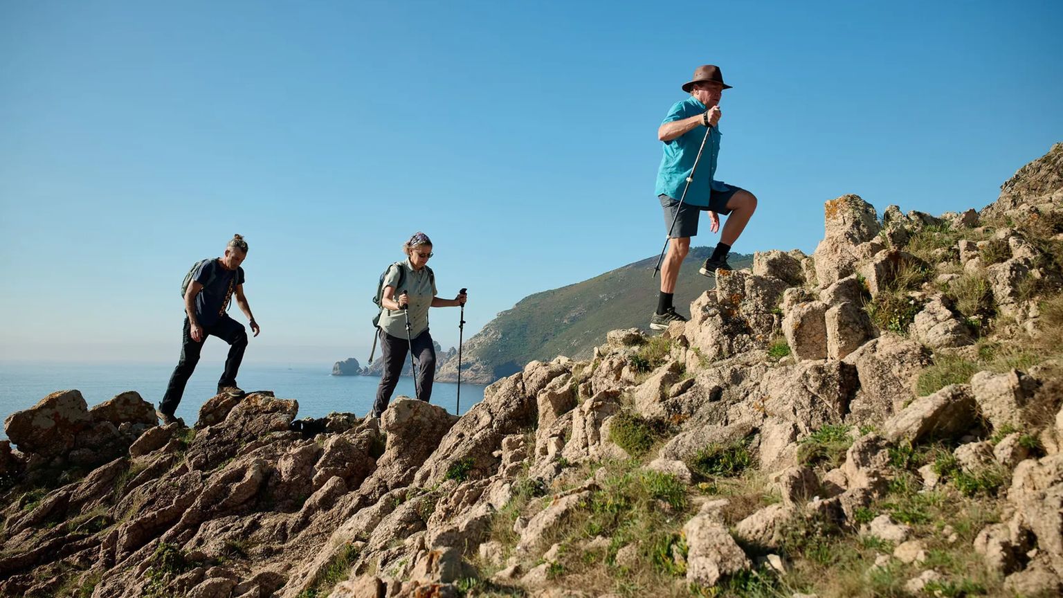 Three hikers with trekking poles climb a rocky hill under a clear blue sky, with coastal scenery in the background.