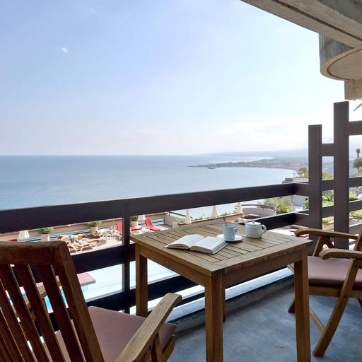 Wooden chairs and a table with an open book and cups on a balcony overlooking a serene ocean view under a clear blue sky.
