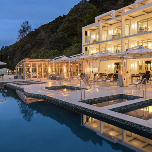Luxury hotel pool area at dusk with illuminated modern building, sun loungers, umbrellas, and a calm, reflective swimming pool.