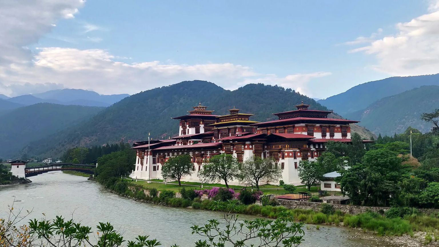 A traditional Bhutanese fortress, or dzong, surrounded by mountains and a river under a partly cloudy sky.