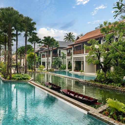 Luxurious resort with villas, palm trees, and a canal-like pool featuring a small wooden boat, under a partly cloudy sky.