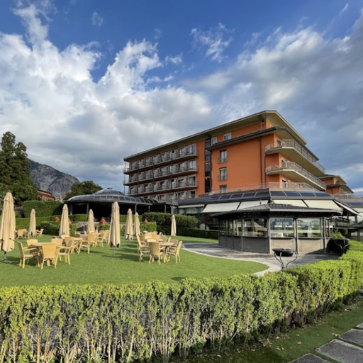 A scenic view of a hotel with outdoor dining area, surrounded by greenery and mountains under a partly cloudy sky.