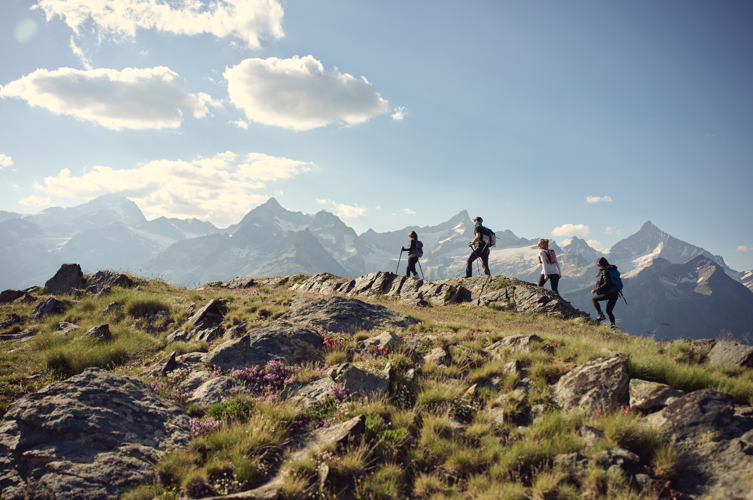 Four hikers walk along a rocky mountain ridge with snow-capped peaks in the background under a partly cloudy sky.