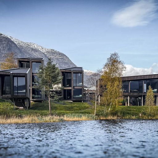 Modern angular buildings by a lake, surrounded by trees and mountains under a blue sky.