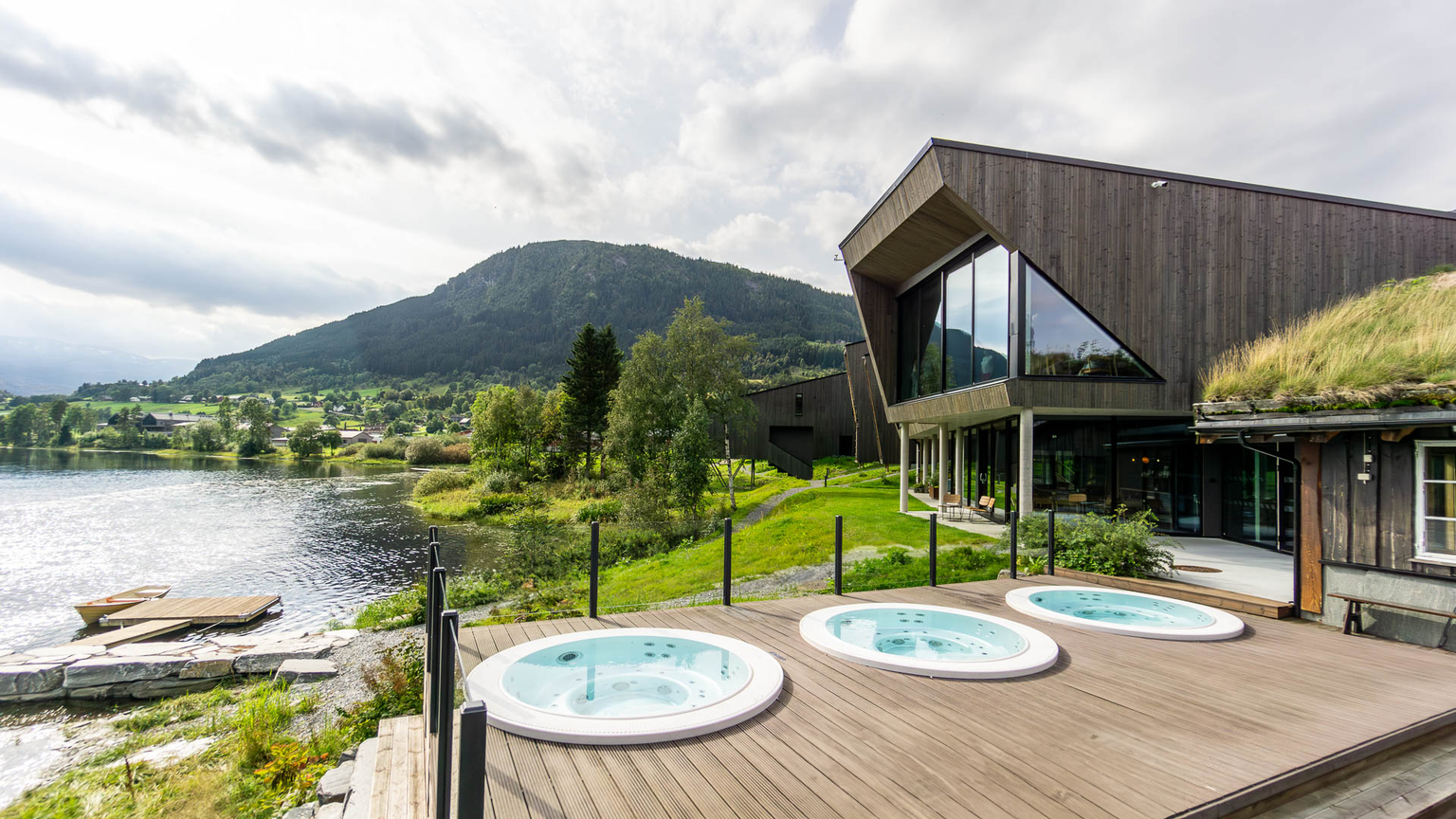 Modern lakeside building with angular design, large windows, and three outdoor hot tubs on a wooden deck, surrounded by lush greenery and mountains.