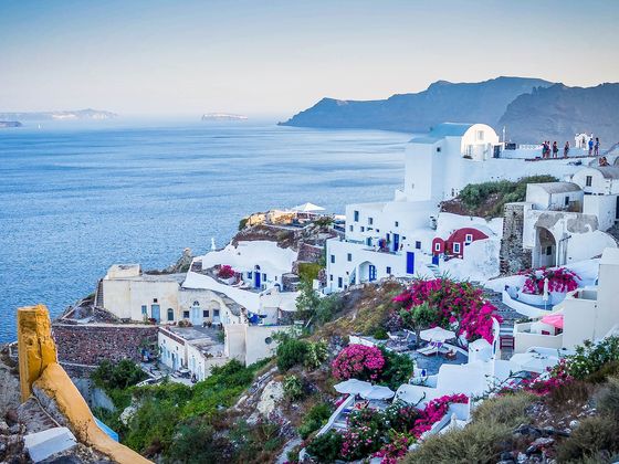 White and blue buildings on a cliffside overlook the sea in Santorini, Greece, with vibrant pink flowers and a mountainous background.